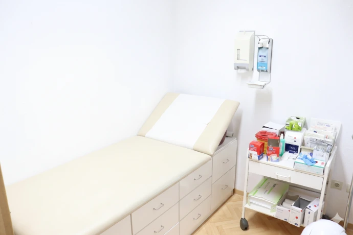 Ein Behandlungszimmer mit Untersuchungsliege, Desinfektionsspender und einem Instrumentenwagen.